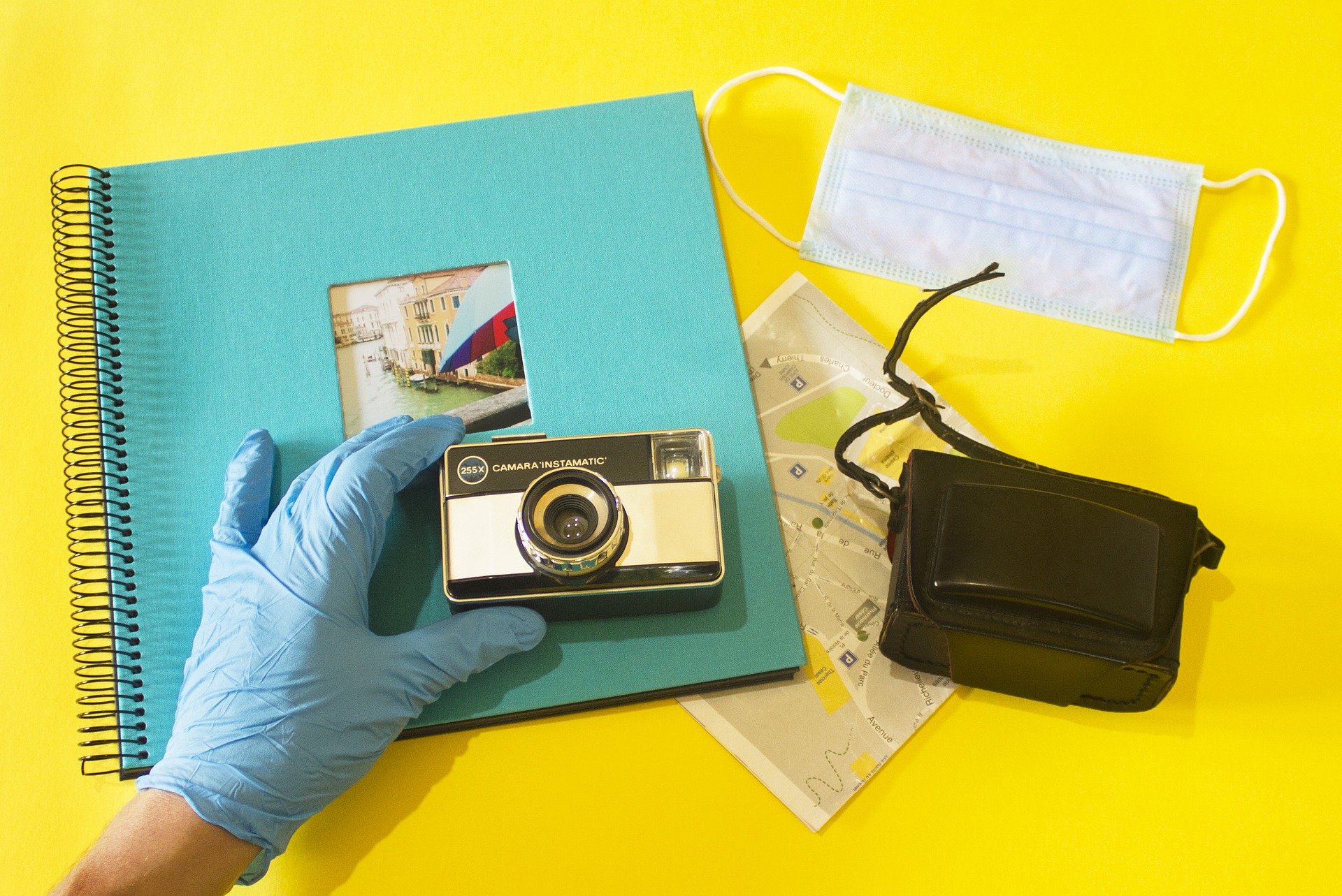 Gloved hand holding camera, mask, map and small black bag to right